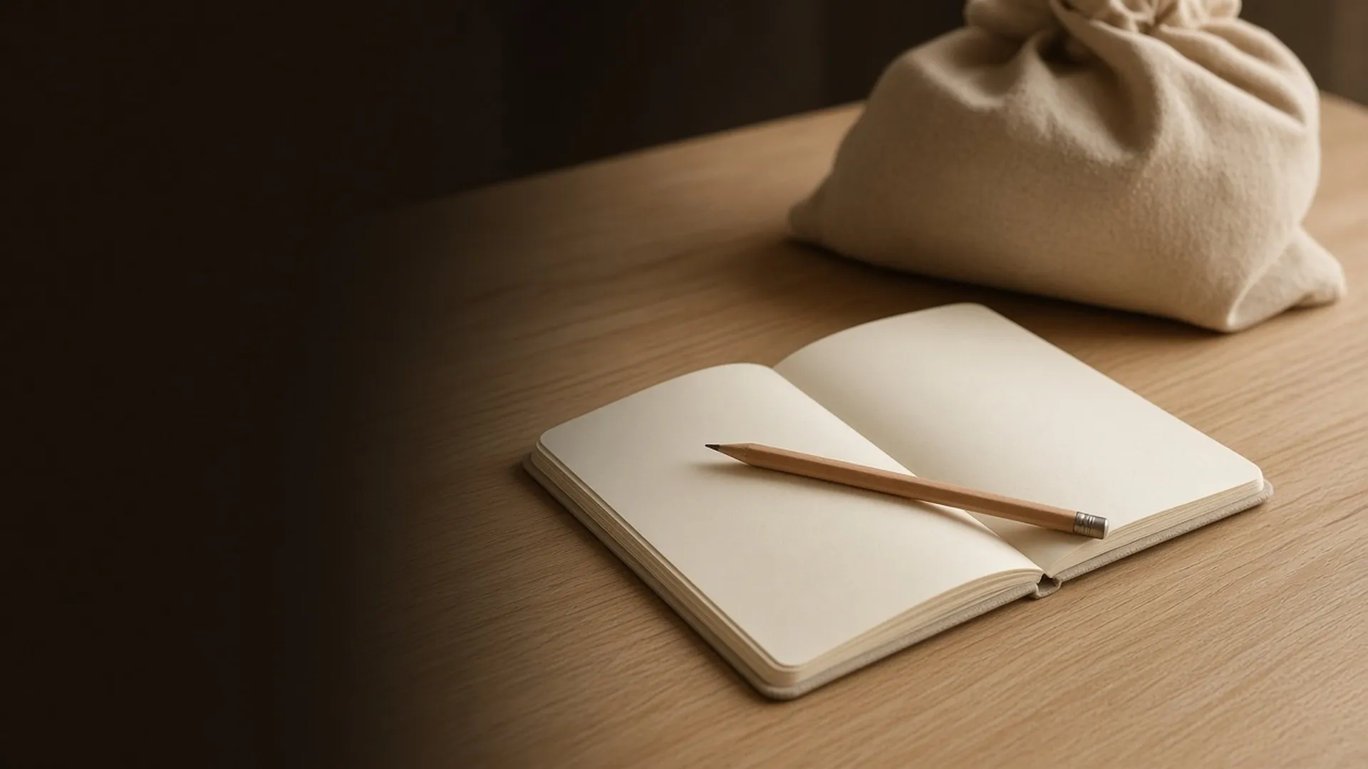 An open notebook and a pencil on an oak table with a bag beyond