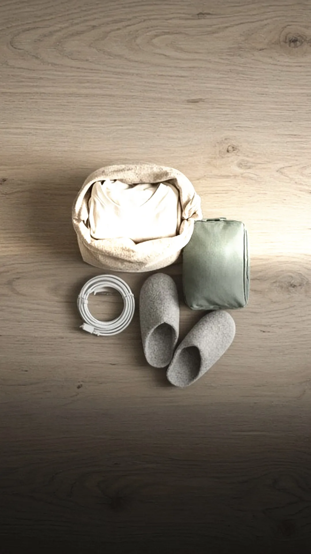 An open bag of folded clothes, a sage pouch, a charger, and grey slippers on oak