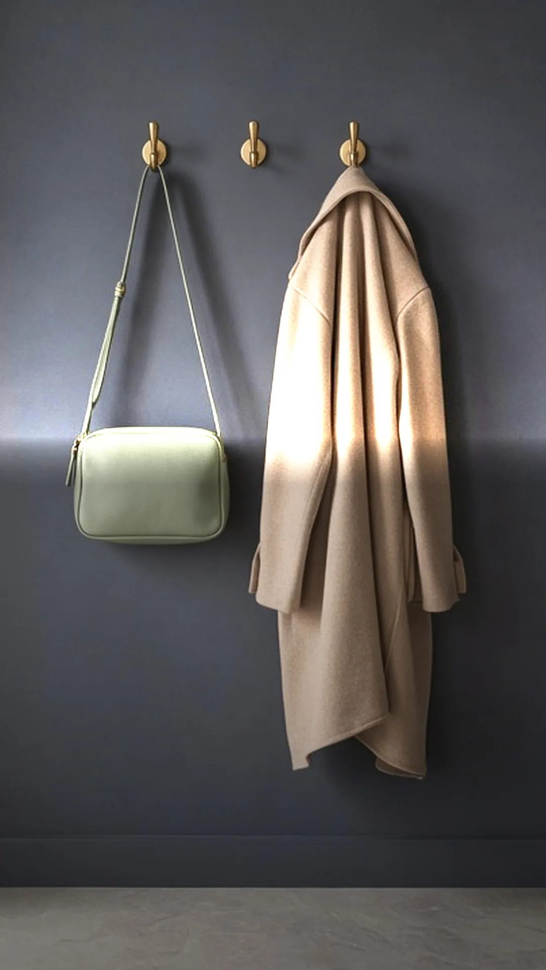 A small sage bag and a tan coat hanging from brass hooks on a dark wall